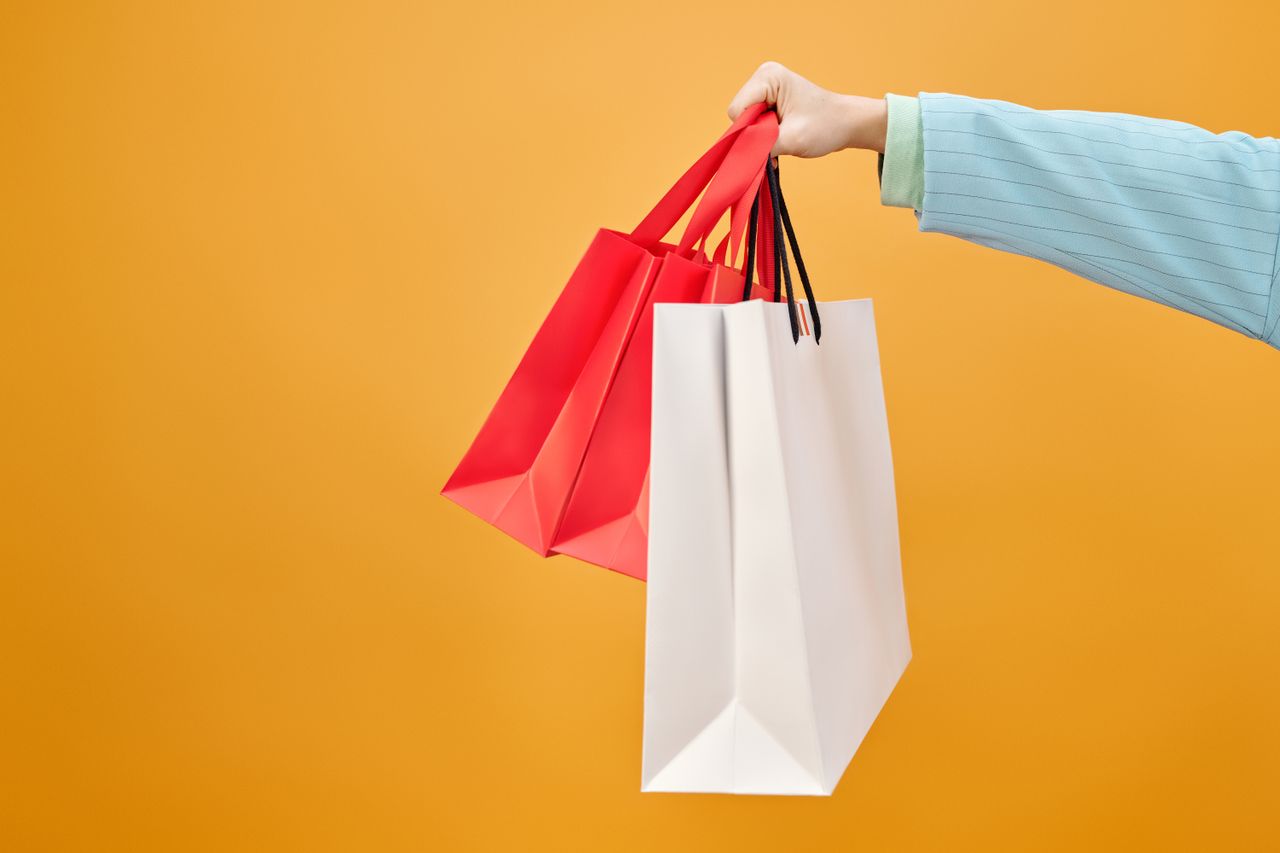 gallery-1 A hand holds colorful shopping bags against a bright yellow backdrop, highlighting retail shopping.